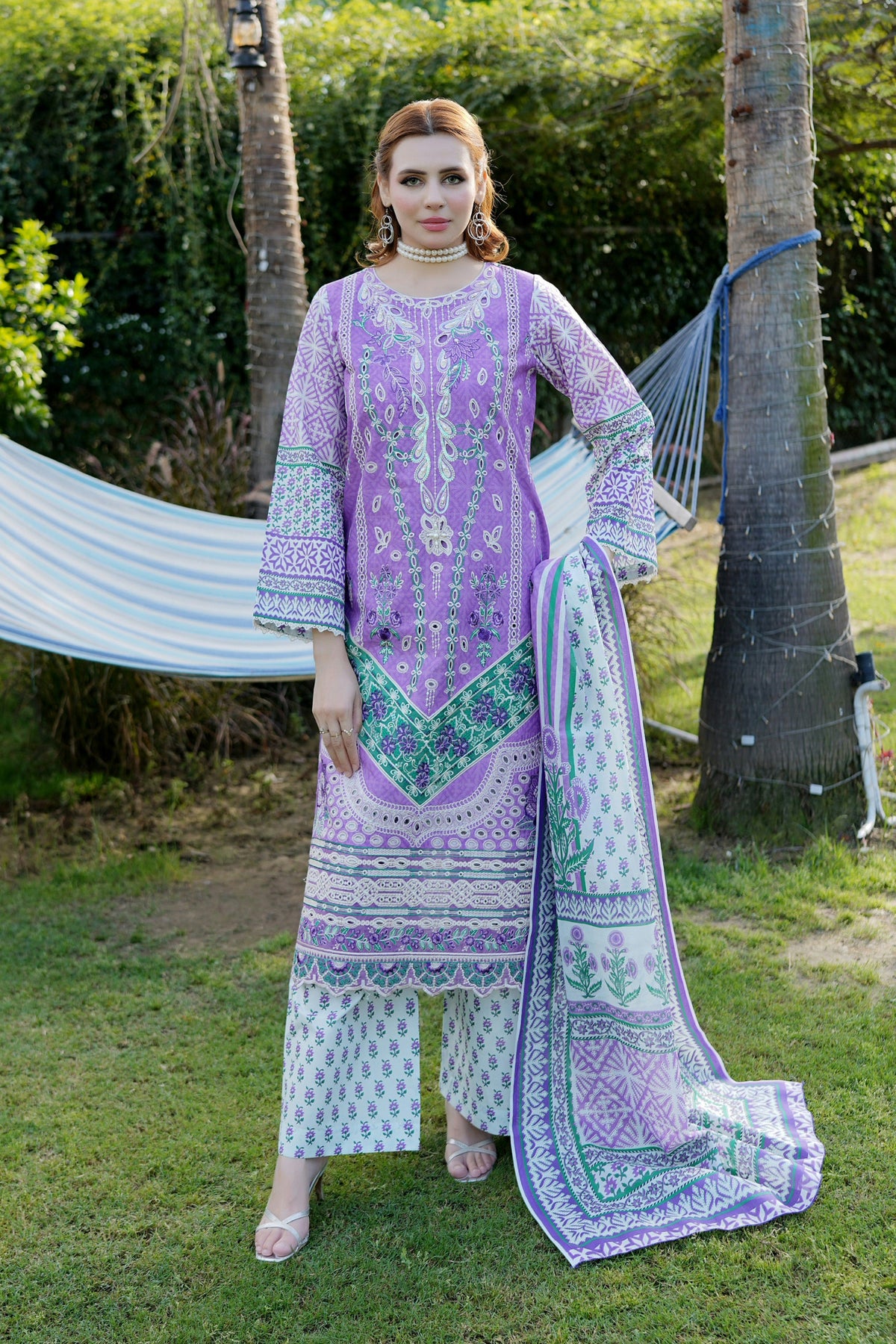 Woman in a purple and white traditional outfit standing outdoors with trees and a hammock in the background.