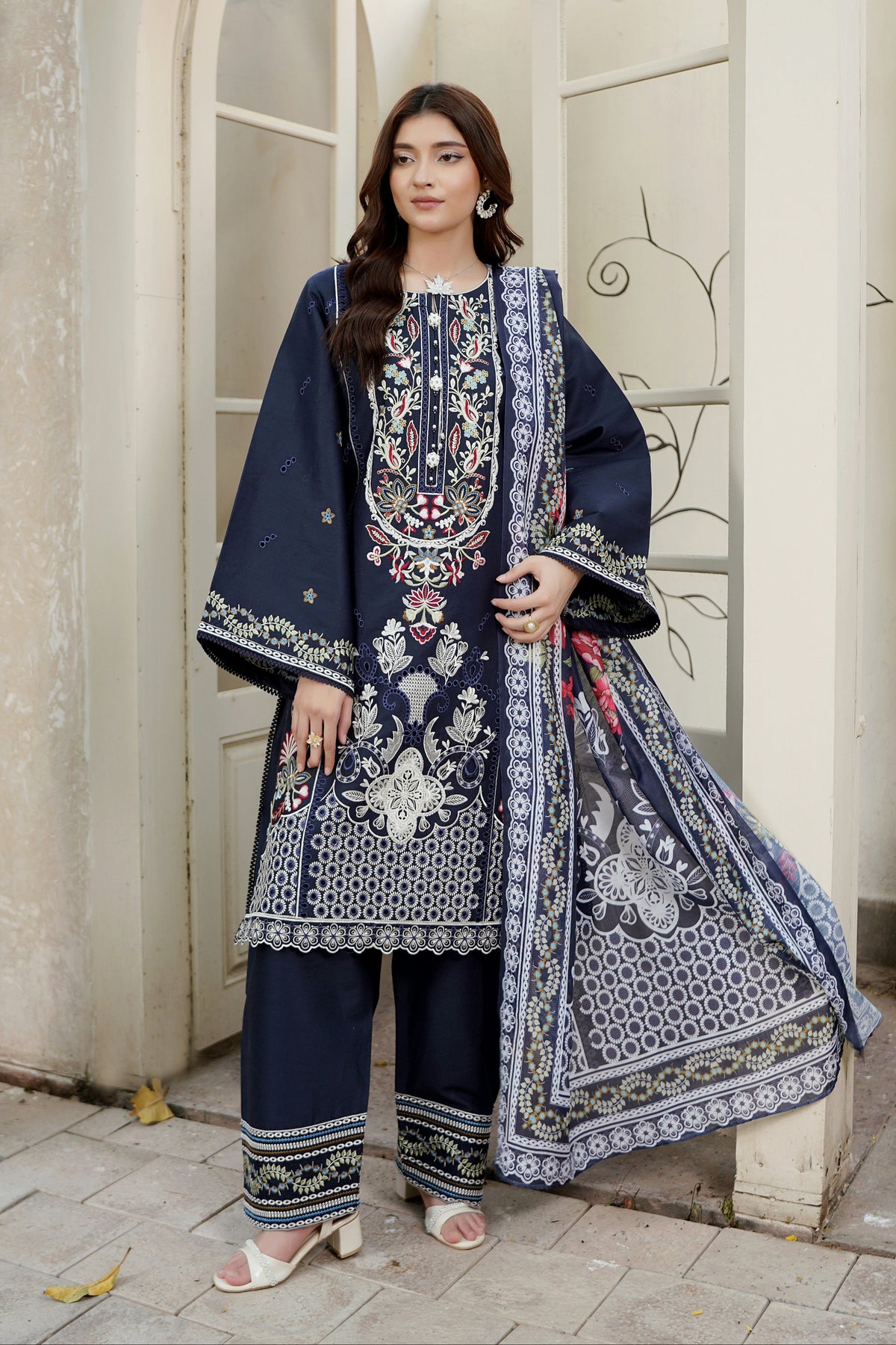 A woman models a navy blue three-piece traditional outfit with intricate white, silver, and red floral embroidery, featuring a long kurta, wide-leg trousers, and a matching dupatta, set against a beige stone archway background.