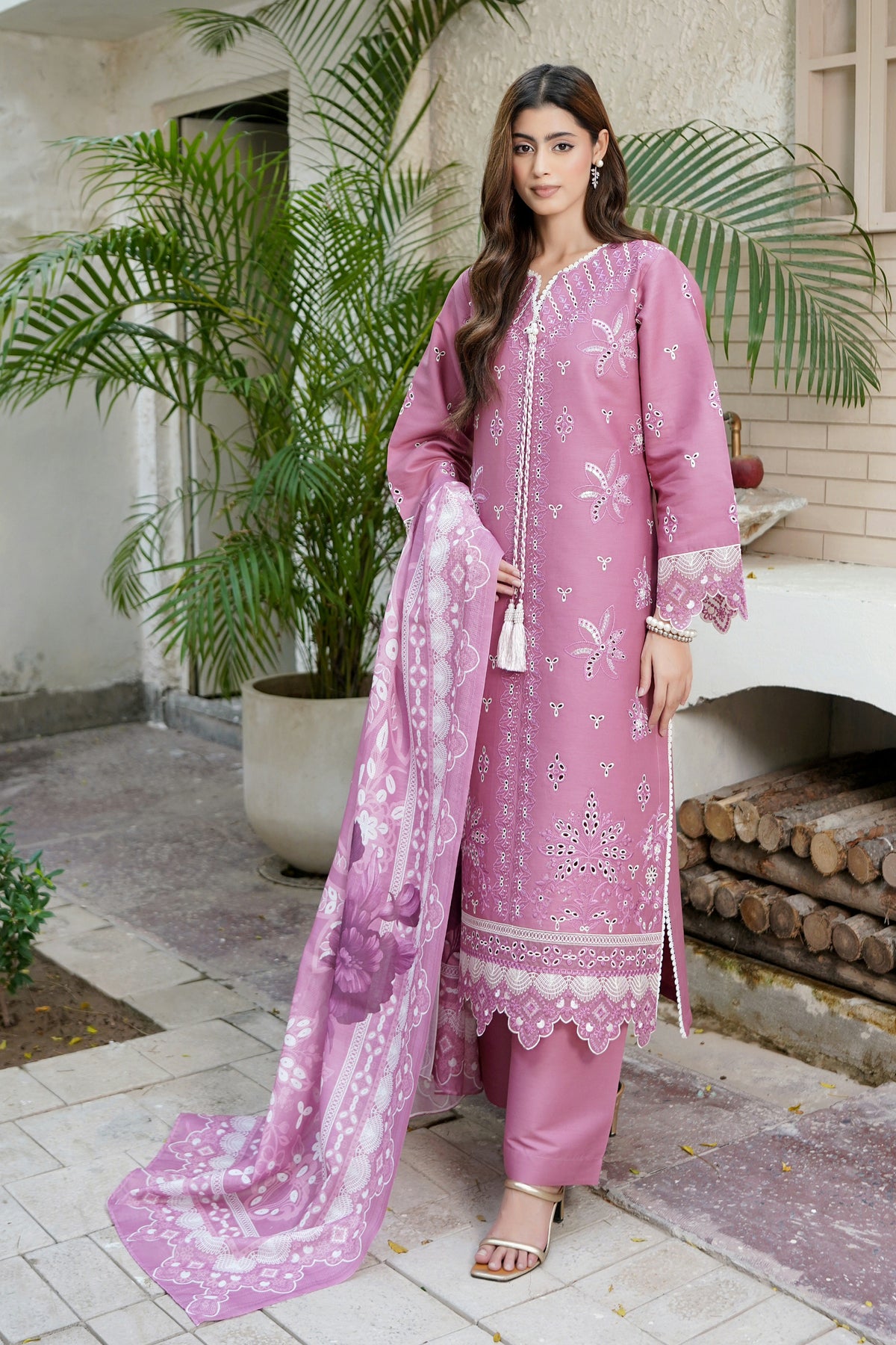 A woman models a three-piece pink ethnic suit with white floral embroidery and a matching dupatta in an outdoor patio setting with potted plants.