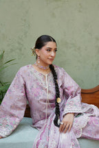 A woman wearing a lavender floral embroidered traditional outfit with long sleeves and a matching skirt, seated against a textured sage green wall.